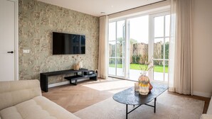A living room with a sofa, a coffee table, a TV, and a sliding glass door leading to a garden. - Family Getaway in Texel (De Koog)
