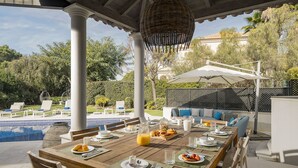 Un espacio comedor al aire libre cubierto con piscina, columnas blancas y un área de descanso con cojines.