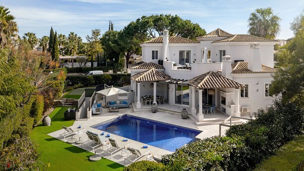 Una villa blanca grande con piscina, mobiliario exterior y un jardín bien cuidado.