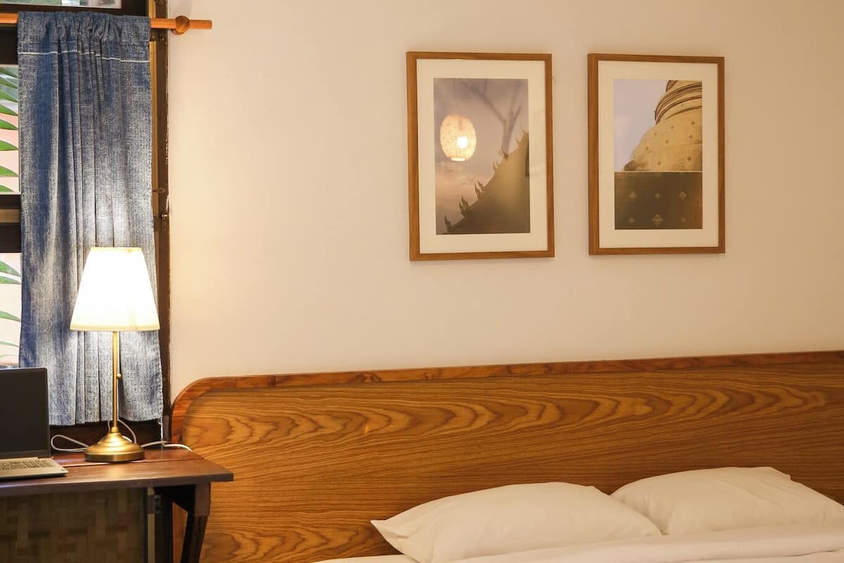A bedroom with a wooden headboard, a bed with white linens, a desk with a lamp, and two framed pictures on the wall.
