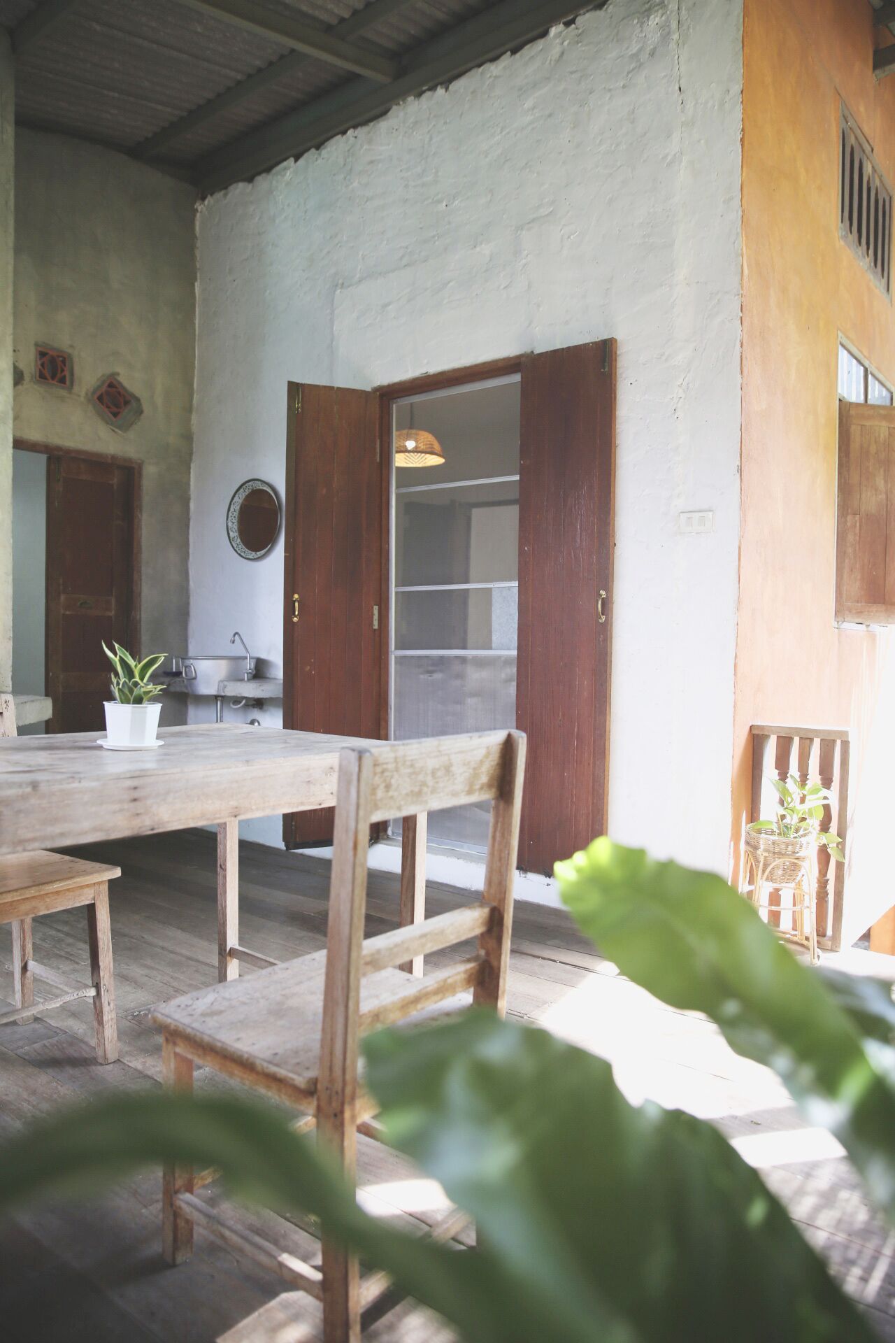 A rustic dining area with a wooden table and chairs, a potted plant, and a view of a kitchen area with a window and a hanging light fixture.
