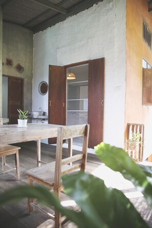 A rustic dining area with a wooden table and chairs, a potted plant, and a view of a kitchen area with a window and a hanging light fixture. - Baannoi Nornmuan Homestay Chiang Mai (Chiang Mai)