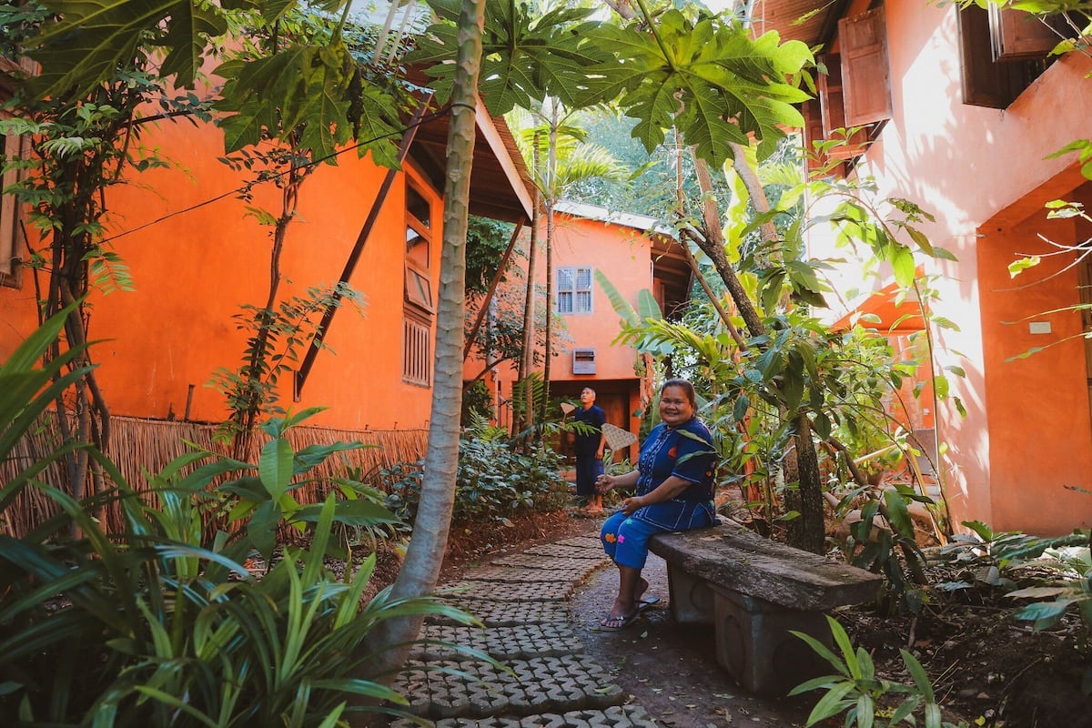 A narrow pathway leading to a building with orange walls and a person sitting on a bench.