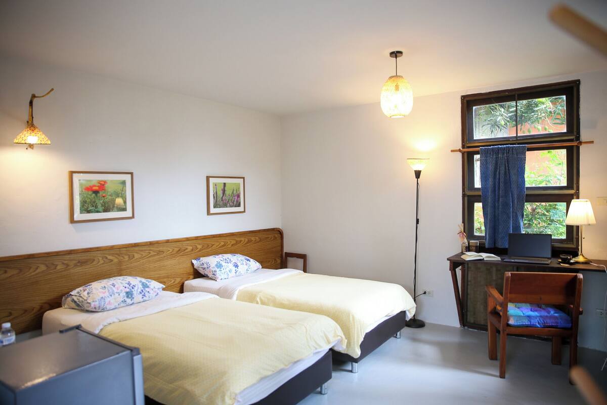 A room with two beds, a desk with a laptop, a chair, and a window with a curtain.