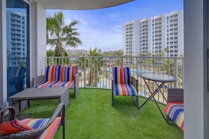 A balcony with wicker furniture, a small table, and a view of palm trees and buildings. - The Palms: 4th flr-Jr Ste, Poolside view | Destin (Destin)