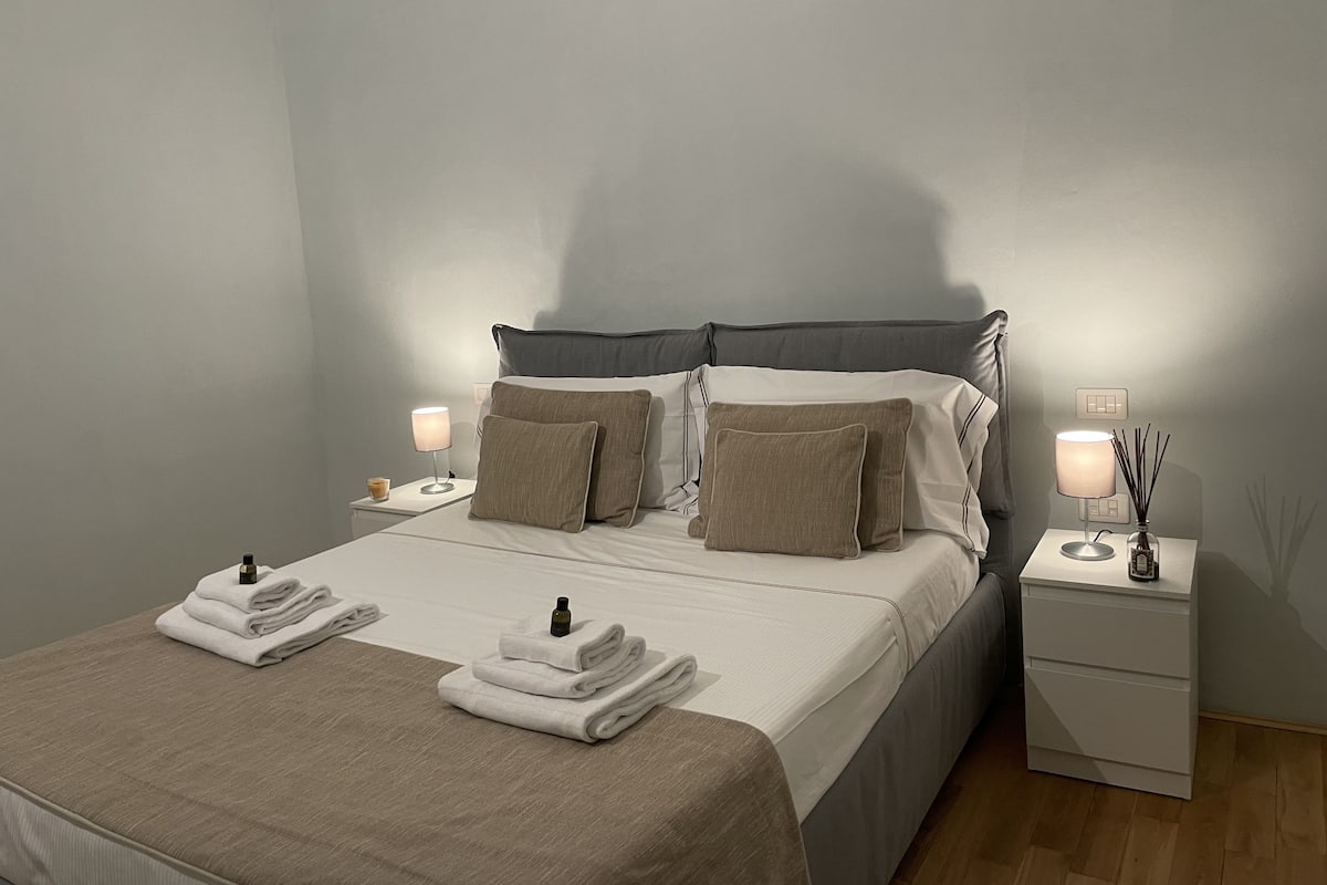 A neatly made bed with pillows, a bedside table with a lamp, and a nightstand with a lamp and a vase.