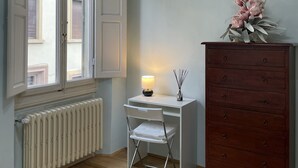Een kamer met een wit bureau, een stoel, een dressoir, een radiator en een raam met geopende luiken.