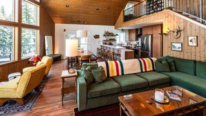 A spacious living area with a green sofa, wooden flooring, and a kitchen in the background. - Summer camp is here at Cle Elum Cabin! Featuring a private hot tub, shuffleboard, games galore, fully stocked kitchen and much more! (Cle Elum)