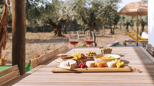 Une table en bois, dressée avec une variété de fromages, de raisins, d’olives et de pain, accompagnée de deux verres de vin rouge, dans un cadre extérieur agrémenté d’oliviers et d’un parasol en chaume.