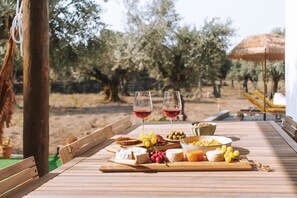 A wooden table set with a variety of cheeses, grapes, olives, and bread, accompanied by two glasses of red wine, in an outdoor setting with olive trees and a thatched umbrella.
