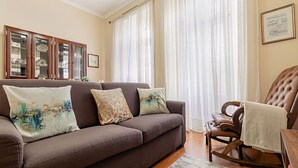 A living room with a brown sofa, three cushions, a wooden rocking chair, and a patterned rug.