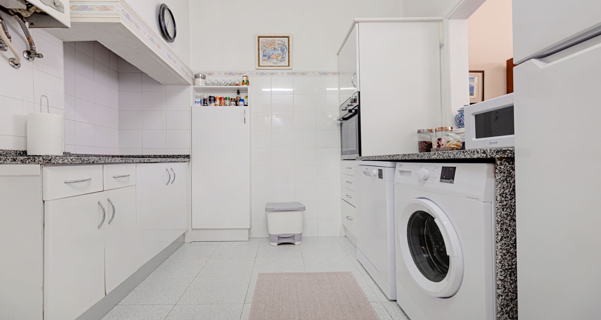A compact kitchen with white cabinets, a built-in washing machine, and a small trash bin.
