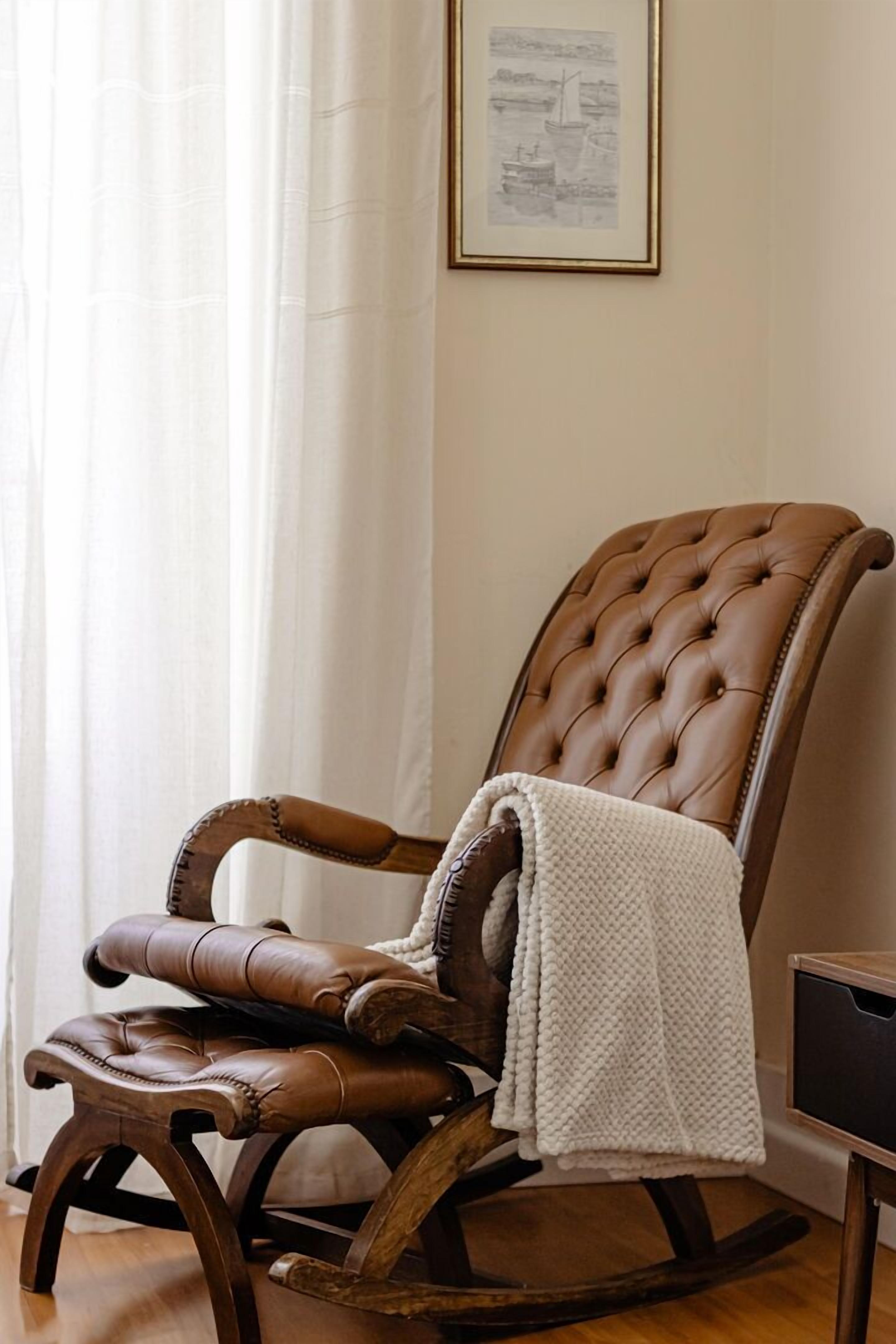 Uma poltrona de couro marrom com um cobertor branco, posicionada perto de uma janela com cortinas brancas.