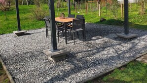 Un'area esterna coperta con tavolo e sedie, circondata da ghiaia ed erba, con alberi e una serra sullo sfondo.