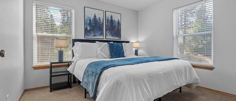 A bedroom with a bed, two nightstands, a window with blinds, and two wall-mounted picture frames.