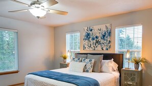 A bedroom with a large bed, a ceiling fan, two bedside tables with lamps, and a floral artwork on the wall. - Rocky Ridge Retreat | Cozy Post Falls Home (Post Falls)