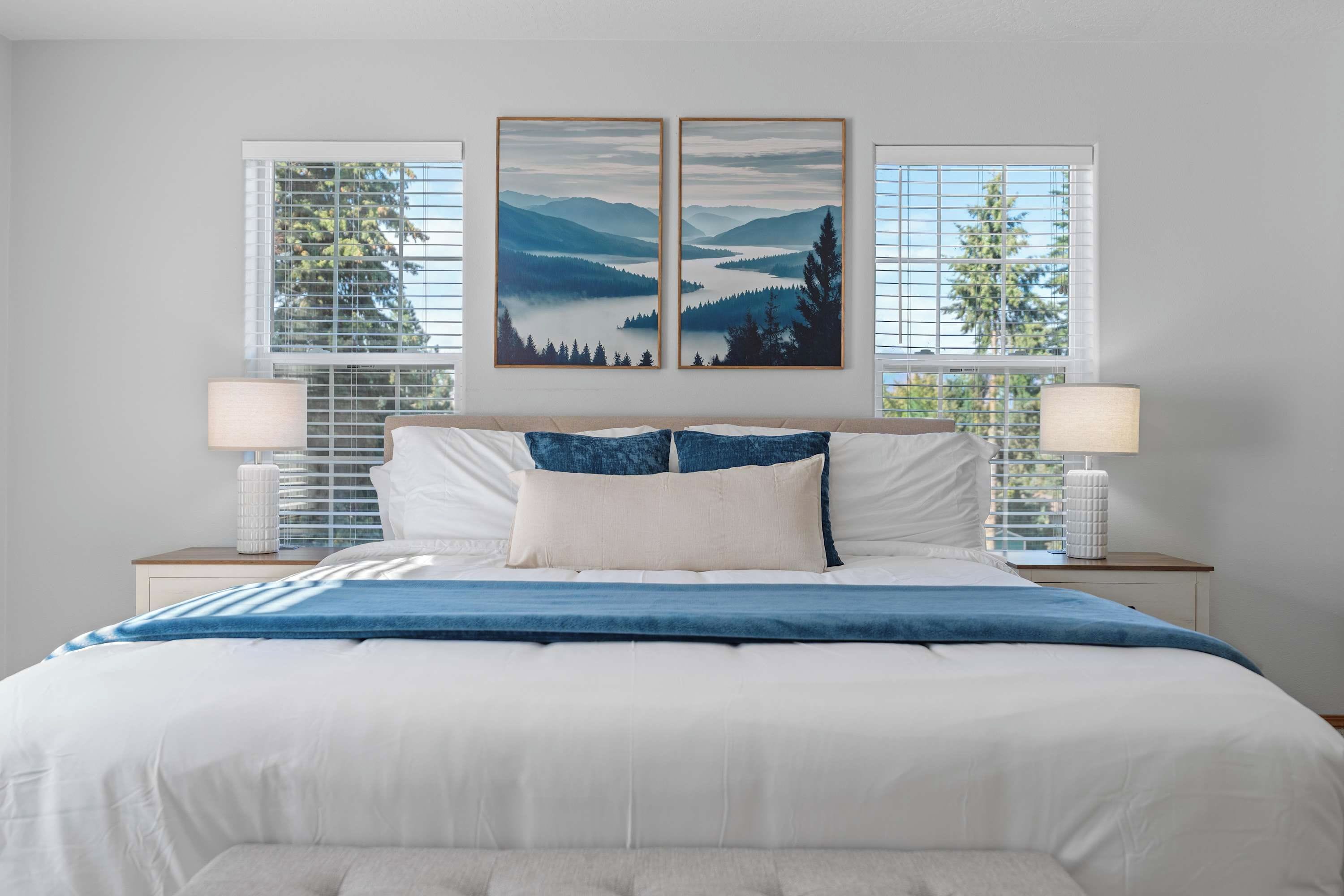 A bedroom with a large bed, two bedside tables, and a view of mountains through the windows.