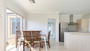 A spacious kitchen with a central island, a dining area with wooden chairs, and a view of the outside through large windows. - Cosy Modern Home close to Fremantle (Coolbellup)