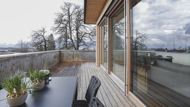 A wooden deck with a table and chairs, potted plants, and a view of trees and mountains through large glass doors. - 100 m2 Penthouse mit 3 Schlafzimmer, Grosser Überdachter Terrasse und Bergblick (Klaus)