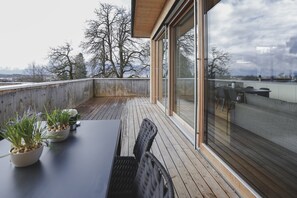 A wooden deck with a table and chairs, potted plants, and a view of trees and mountains through large glass doors.