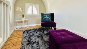 A room with a wooden floor, a black and white rug, a purple ottoman, a teal cushion with text, a small table with a lamp and plant, and a window with white shutters.