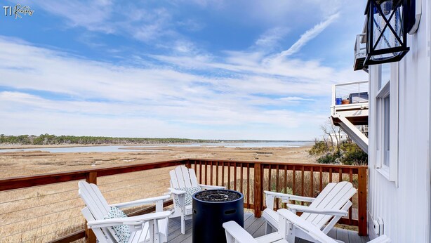 A deck with white Adirondack chairs, a black trash can, and a view of a beach and marshland. - Otter Space | Waterfront | Dog Friendly | Hot Tub (Surf City)