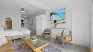 A modern living room with a bed, two armchairs, a coffee table, and a TV mounted on the wall displaying a beach scene. - Sandy Toes Studio: Your Beachfront Getaway Awaits (Fort Walton Beach)