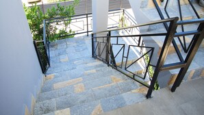 A staircase with stone steps and metal railings, leading to an outdoor area with greenery and a building in the background.