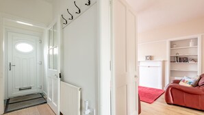 Un pasillo con una puerta blanca, un radiador, un perchero y una alfombra roja que conduce a una habitación con un sofá rojo y una estantería.