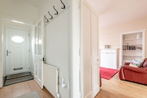 A hallway with a white door, a radiator, and a red rug leading to a room with a red sofa and a bookshelf. - Boase Avenue (No 24) | St Andrews (St Andrews)