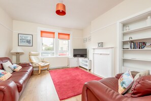A living room with a red carpet, leather sofas, a wooden chair, a TV, a bookshelf, and a window with orange blinds. - Boase Avenue (No 24) | St Andrews (St Andrews)