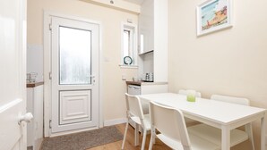 Una habitación pequeña y limpia con una puerta blanca, una mesa blanca con sillas, una pequeña zona de cocina y un cuadro en la pared.