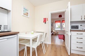 A compact kitchen with white appliances, a dining area with a white table and chairs, and a view into a living space with a red rug. - Boase Avenue (No 24) | St Andrews (St Andrews)