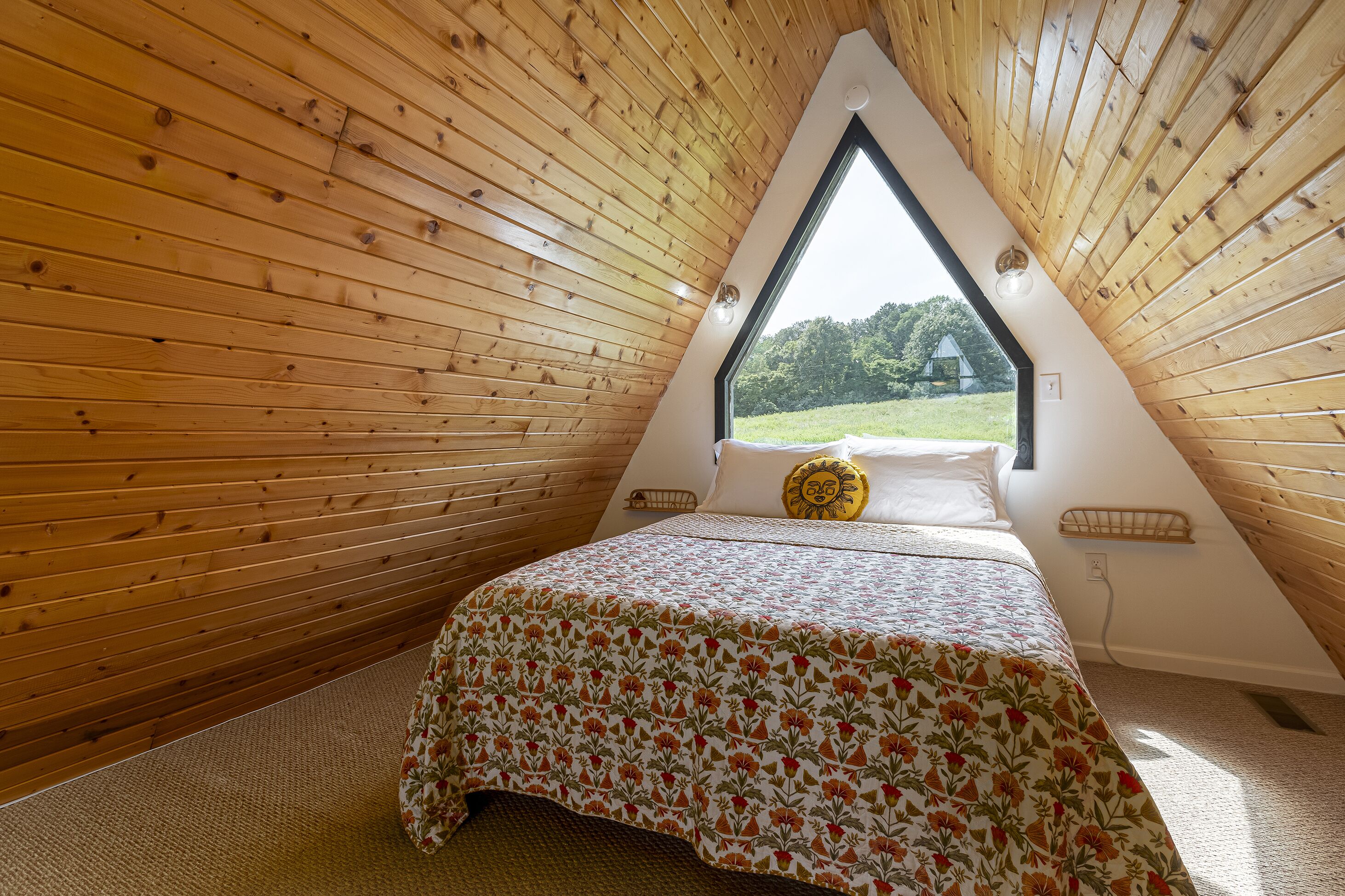 A triangular attic bedroom with wooden walls, a bed with a floral bedspread, and a window offering a view of greenery.