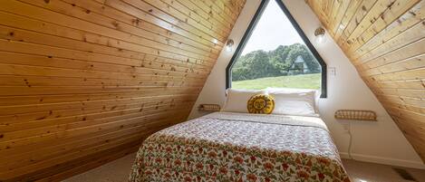 A triangular attic bedroom with wooden walls, a bed with a floral bedspread, and a window offering a view of greenery.