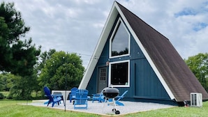 Un edificio a forma di triangolo con un'ampia finestra e un portone, immerso in un prato verde con sedie blu e un barbecue.