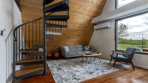 Una stanza in stile loft con soffitto in legno, una scala a chiocciola, un divano, un tavolino da caffè e un'ampia finestra vista sul verde.