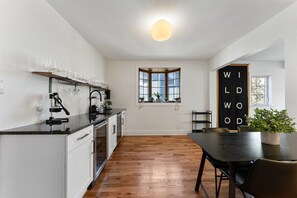 Cozinha moderna com armários brancos, bancada preta e piso de madeira. Há uma janela com vista para o exterior, e uma placa preta com letras brancas na parede.