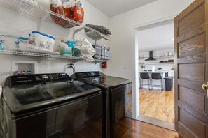 Een wasruimte met twee voorladers wasmachines en drogers, planken erboven met diverse huishoudartikelen, en uitzicht op een keuken met barstoelen.