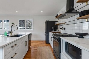 A modern kitchen with white cabinetry, a central island, a stove, and a refrigerator. - Spacious 7BR Beach House with Arcade and Porch! (Wildwood)