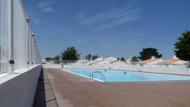 Une piscine sous un ciel bleu azur, un bâtiment blanc avec une rambarde en métal, et quelques arbres en arrière-plan.
