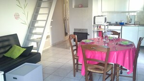 Un coin repas avec une nappe rose, des chaises en bois et une table à manger. Un canapé blanc avec un coussin vert est placé contre le mur. Dans la cuisine, on trouve un meuble blanc et un four à micro-ondes.