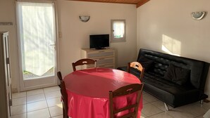Une pièce avec un plafond en bois, une table à manger recouverte d’une nappe rouge, un canapé en cuir noir, un téléviseur posé sur un meuble en bois et une fenêtre donnant sur un paysage verdoyant.