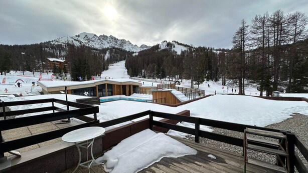 Estación de esquí con terraza de madera, piscina y montañas al fondo.