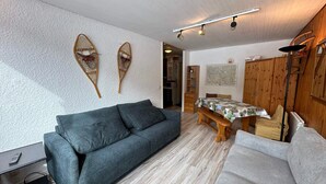 Una sala de estar acogedora con un sofá gris, una mesa de comedor de madera con mantel estampado y un estante con adornos.