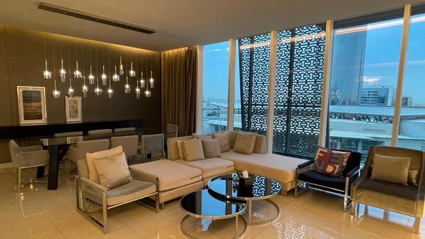 Ein modernes Wohnzimmer mit einer Couch, Sesseln, einem Couchtisch und einem Essbereich mit Stühlen und einem Tisch. Große Fenster bieten Blick auf eine Stadtlandschaft.