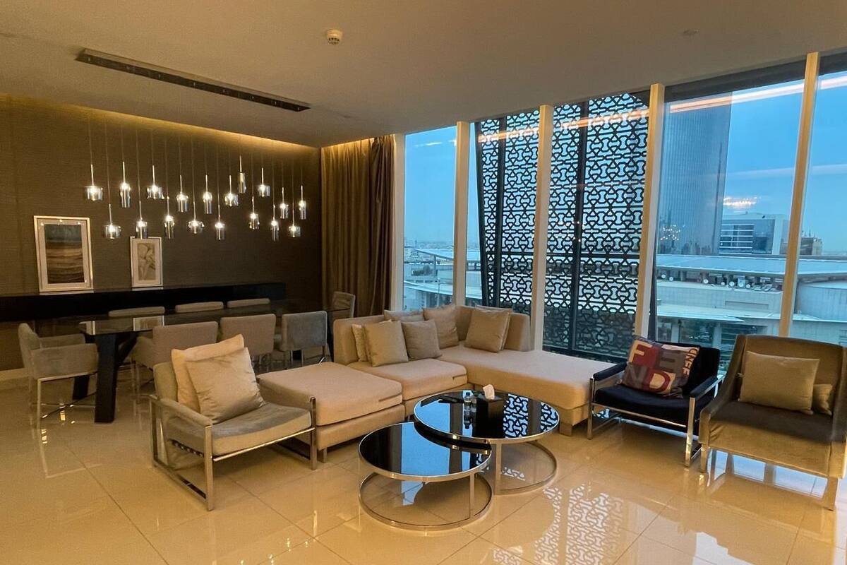 Ein modernes Wohnzimmer mit einer Couch, Sesseln, einem Couchtisch und einem Essbereich mit Stühlen und einem Tisch. Große Fenster bieten Blick auf eine Stadtlandschaft.