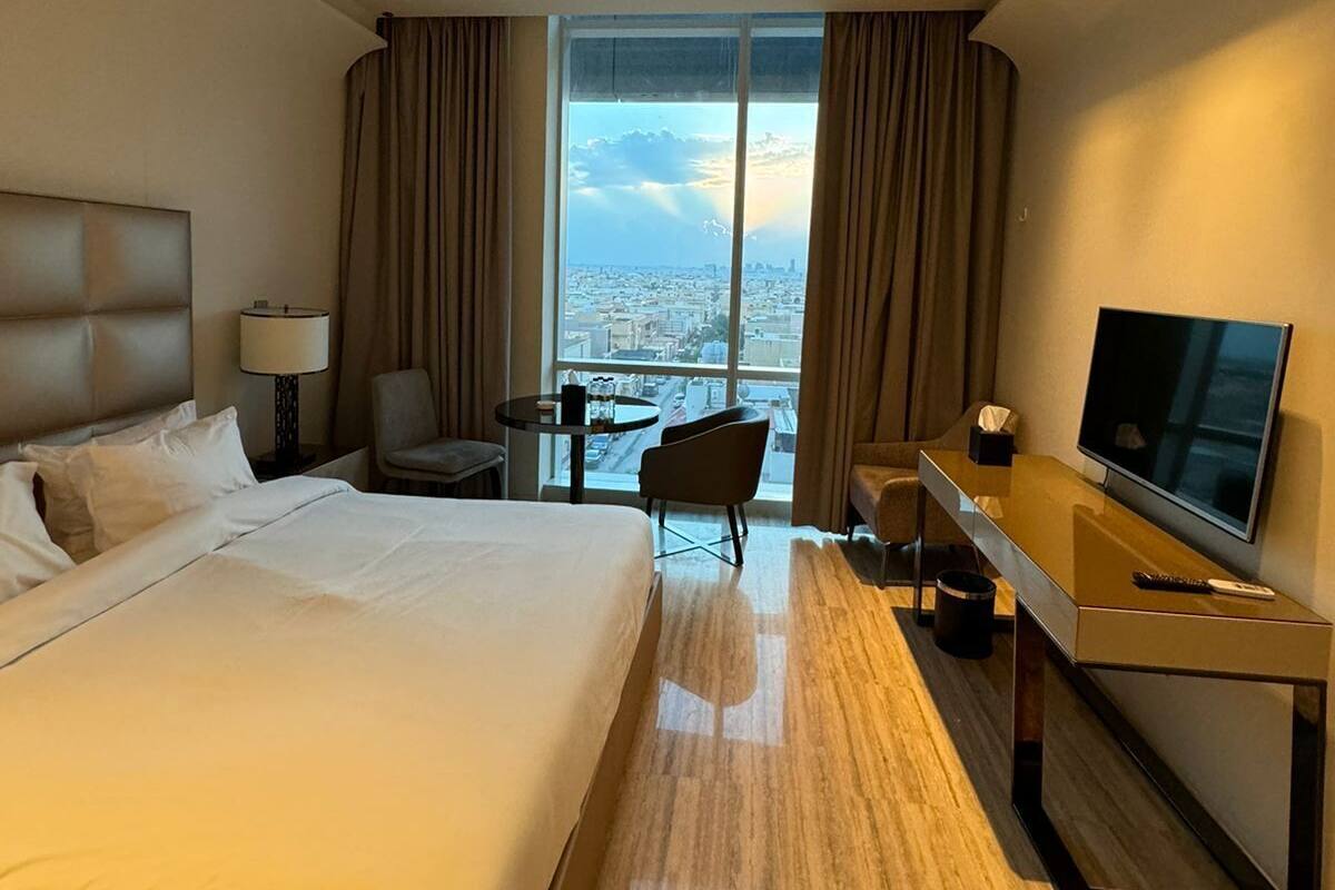 Ein Hotelzimmer mit einem großen Bett, einem Schreibtisch mit Fernseher, einem Sessel und Blick auf die Stadt durch große Fenster.