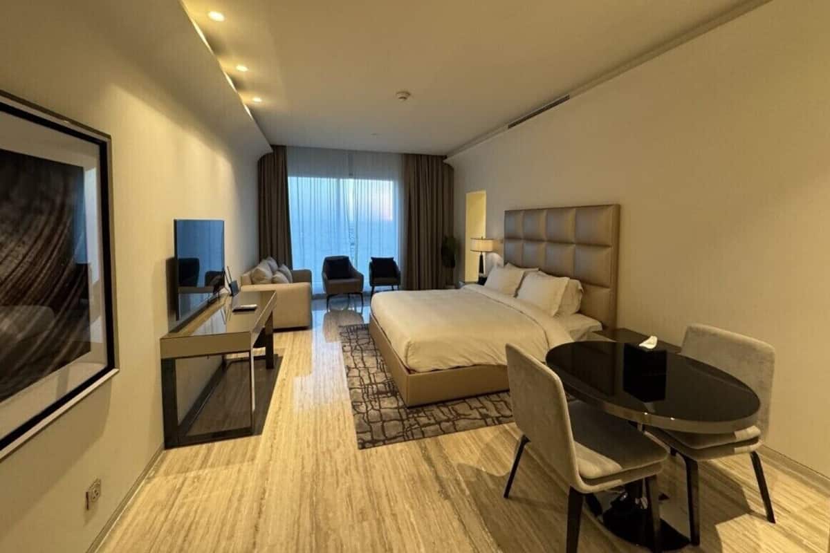 Ein modernes Hotelzimmer mit einem großen Bett, einem Schreibtisch mit Stuhl, einem Esstisch mit Stühlen und einem großen Fenster mit Vorhängen.