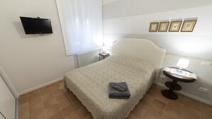 A bedroom with a bed, a TV mounted on the wall, a window with curtains, a bedside table with a lamp, and framed pictures on the wall. - Family room with 2 double or twin rooms near the port and the Venice district. (Livorno)